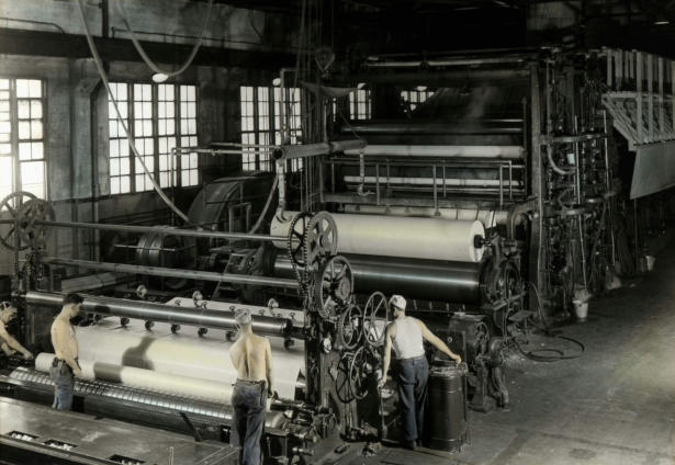 Paper production factory with gigantic machine with half-naked male workers working aside, from picture decades ago